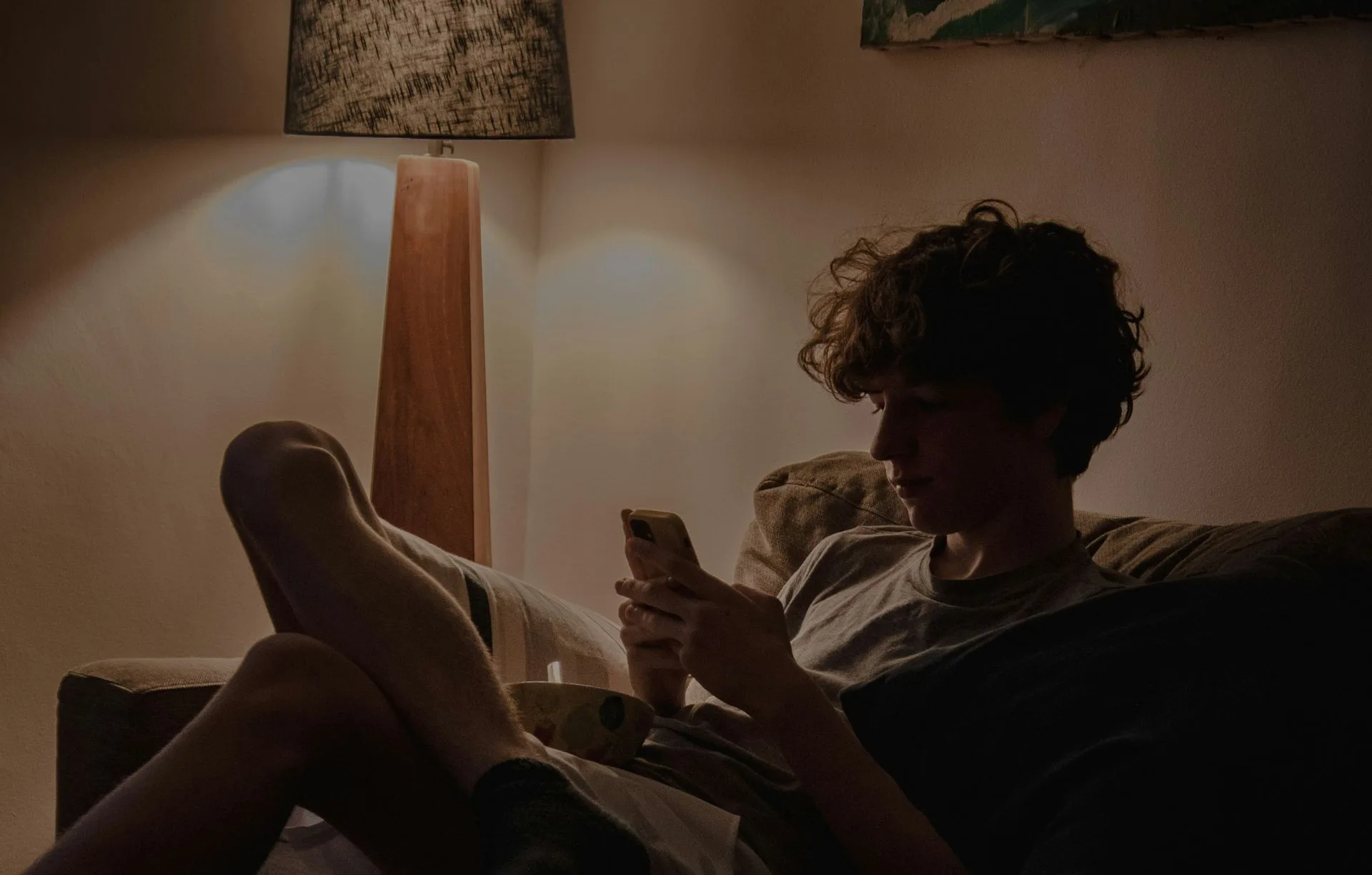 Someone resting on a sofa in the evening, feet up, phone in hand, in the warm light of a floor lamp